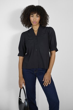 Blusa nera a maniche corte con vestibilità comoda, dotata di colletto arricciato e bottoni sul davanti. Indossata con jeans blu scuro e una borsa nera.