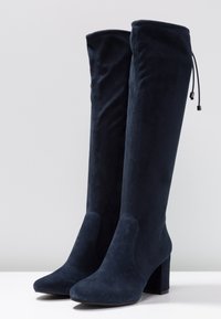 Mörkblå mockastövlar som går över knät med spetsig tå, klackigt stycke och justerbar knytning upptill. Slät textur och åtsittande design.