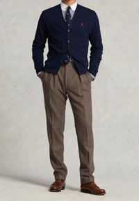 Marinblå ulkardigan, knappad, med rött logotyp. Vit skjorta med blå ränder, brunrutiga byxor, bruna läderskor.