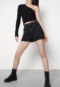  Schwarzes geripptes Crop-Top mit einem Schulterträger. Hoch taillierte schwarze Jeansshorts mit umgeschlagenen Säumen. Modell trägt schwarze Schnürstiefel im Combat-Stil.