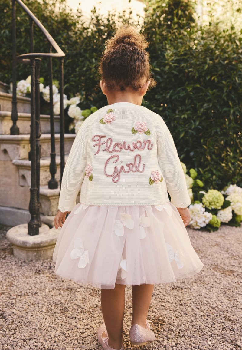 Jong kind in een witte trui geborduurd met "Flower Girl" en bloemdecoraties, met een tule rok en glinsterende schoenen, staand buiten.