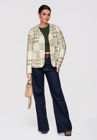 Femme portant un cardigan vert à motifs, un crop top vert foncé, un jean large foncé, une ceinture marron, des chaussures marron, tenant un sac à main beige.
