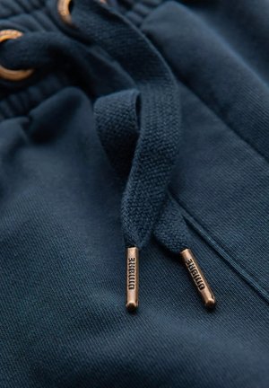 Gros plan sur un sweat à capuche bleu marine avec un cordon de serrage ; présente un tissu texturé et des embouts métalliques en bronze gravés du nom de la marque.