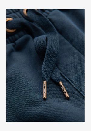 Lähiakuva tummansinisestä hupparista, jossa on nyöri; ominaisuuksiin kuuluu teksturoidusta kankaasta valmistettu malli ja pronssiset metalliset päätylangat, joissa on painettu brändin nimi.