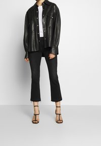 Personne portant un jean flare noir raccourci, une chemise en cuir noire boutonnée par-dessus un haut blanc, et des sandales à talons hauts noires à lanières.