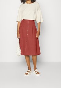 Femme portant un haut beige à manches courtes, une jupe midi couleur rouille avec des boutons sur le devant, des sandales blanches et tenant un sac en filet blanc.