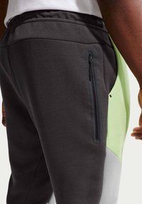 Vue latérale d'une personne portant un pantalon noir, vert clair et gris avec une poche verticale zippée noire sur la cuisse.