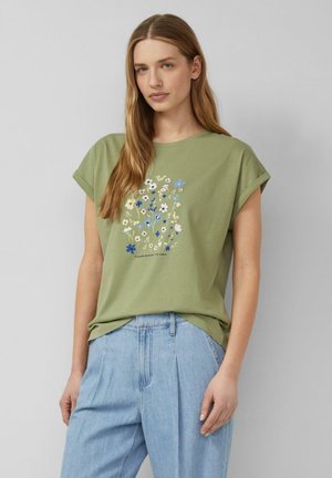 Vrouw met een lichtgroen T-shirt met een meerkleurige bloemenprint en opgerolde mouwen, gecombineerd met lichtblauwe hoge taille jeans.