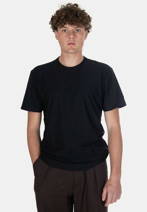 Giovane uomo con capelli ricci che indossa una maglietta nera semplice a maniche corte e pantaloni marrone scuro, in piedi su uno sfondo bianco.