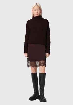 Vrouw met lang blond haar, gekleed in een donker geribd truitje, rok met kantafwerking en zwarte kniehoge laarzen, staand tegen een effen achtergrond.