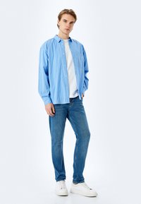 Camisa de botones a rayas azul claro, camiseta de cuello redondo blanca por debajo, jeans azules y zapatillas blancas. Ajuste casual con mangas holgadas.