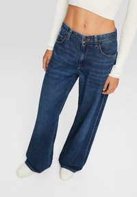 Jeans blu a gamba larga in denim, con vita alta, leggero effetto scolorito sui fianchi e cinque tasche. Indossati con una maglietta bianca e sneakers.