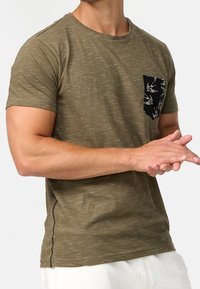 Camiseta de manga corta verde oliva hecha de tejido texturizado, con un bolsillo negro que presenta un patrón de pájaros en blanco y detalles de costura.
