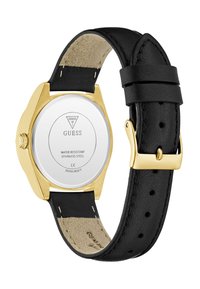 Aukščiau auksinės spalvos "Guess" laikrodžio su juodu odiniu dirželiu atvirkštinė pusė, rodanti nerūdijančio plieno dugną, ant kurio išgraviruoti prekės ženklas ir vandeniui atsparumo detalės.
