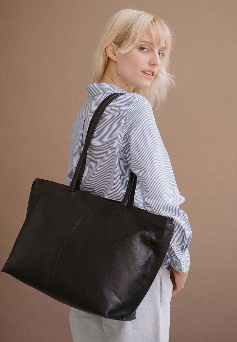 Borsa a tracolla nera in pelle con due manici. Texture liscia, forma rettangolare e delicati dettagli delle cuciture. Indossata con una maglietta a righe.