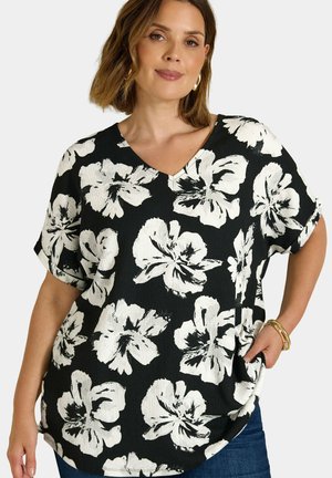 Mujer con blusa negra de manga corta y escote en V con gran estampado floral blanco, vaqueros azules, pendientes de aro y pulseras doradas, con la mano en el bolsillo.