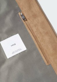 Tissu gris avec étiquette de marque MNG blanche cousue à l’intérieur et une poche zippée en daim beige au-dessus à l’intérieur du vêtement.