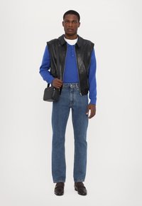 Black leather vest over a blue long-sleeve shirt, paired with light blue jeans and dark loafers, carrying a small black handbag.