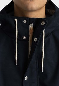 Navy blue jacket with a drawstring hood, metal snaps, and a zippered front. Features a textured fabric and contrast stitching.