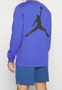 Lila långärmad tröja med en stor svart silhuett av en hoppande figur på baksidan. Bärs med blå meshshorts.