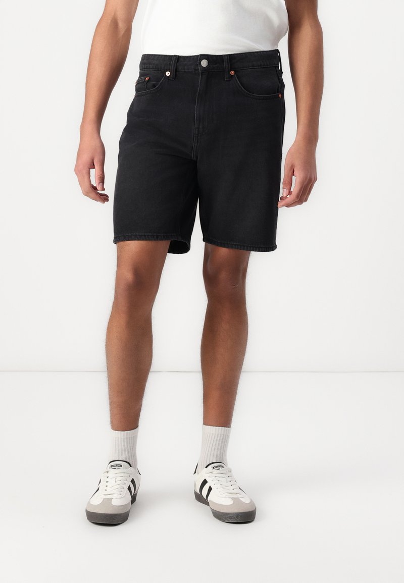 Shorts din denim negru cu un design clasic cu cinci buzunare, prezentând o închidere cu nasture și detalii de cusătură subtile. Stilizați cu pantofi sport albi.
