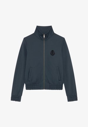 Chaqueta navy con cuello alto, cierre completo, dos bolsillos laterales, puños elásticos y un escudo texturizado negro en el pecho.