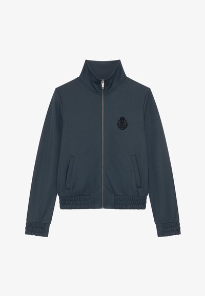 Giacca navy con colletto alto, zip completa, due tasche laterali, polsini elasticizzati e un distintivo nero testurizzato sul petto.