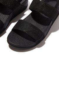 Sandali neri con suola in gomma testurizzata, caratterizzati da due ampie cinghie in tessuto. Il materiale presenta una leggera lucentezza, che arricchisce il design complessivo.