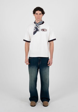 Mann trägt weißes Kurzarmhemd mit PMC-Logo, blau kariertes Halstuch, ausgewaschene blaue Jeans und braune Wildlederschuhe, steht vor weißem Hintergrund.