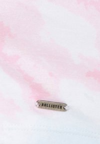 Camiseta de algodón ligera con un diseño de tie-dye en rosa y blanco. Incluye una etiqueta de metal plateado "HOLLISTER" cerca del dobladillo.