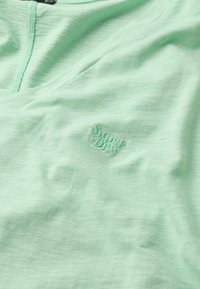 Camiseta de color verde menta clara hecha de una tela suave y ligera; presenta un sutil patrón texturizado y un logo emborrachado de "Superdry" en el frente.