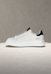 Λευκό sneaker χαμηλού τύπου με μαύρη φτέρνα, κορδόνια και χρυσό λογότυπο "Just Cavalli" στο πλάι, τοποθετημένο σε ουδέτερο φόντο.
