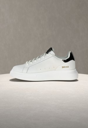 Bela nizka superga s črnim jezičkom na peti, vezalkami in zlatim logotipom "Just Cavalli" na strani, postavljena na nevtralen ozadje.