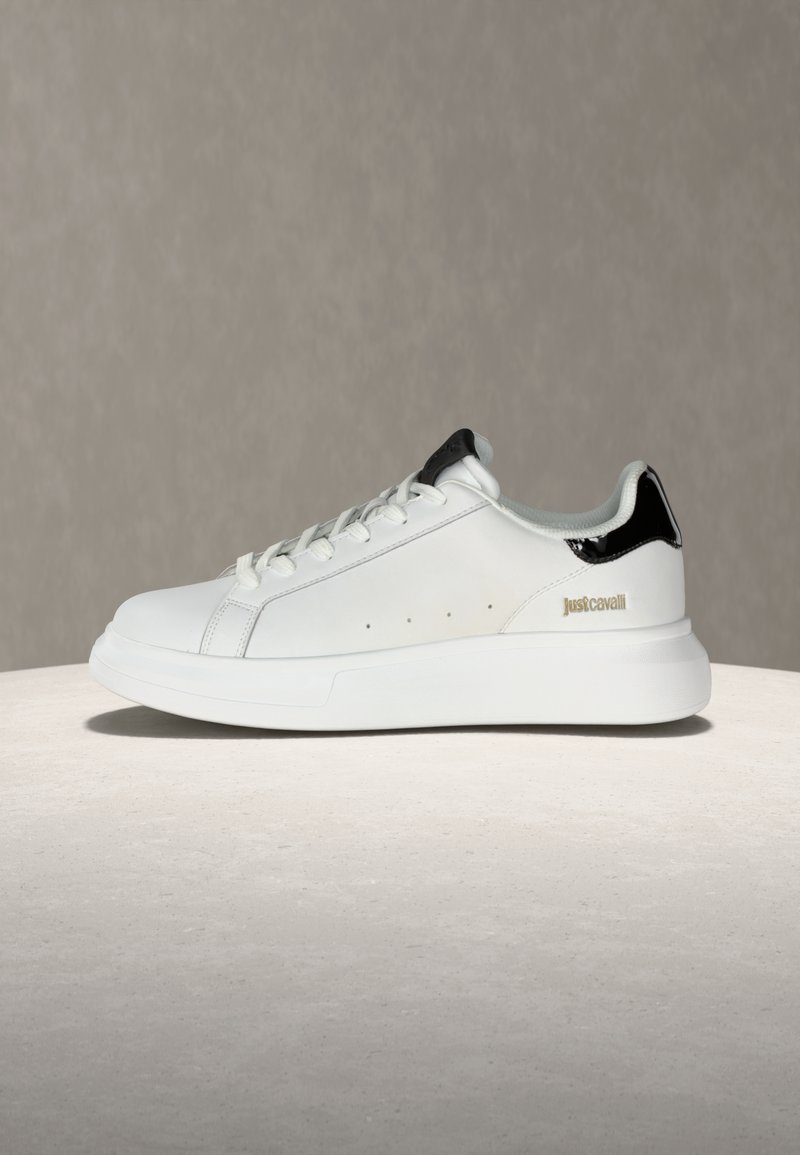 Λευκό sneaker χαμηλού τύπου με μαύρη φτέρνα, κορδόνια και χρυσό λογότυπο "Just Cavalli" στο πλάι, τοποθετημένο σε ουδέτερο φόντο.