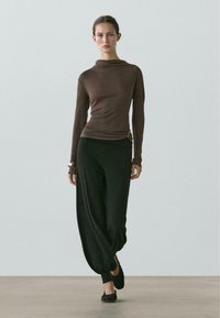 Braunes Langarmshirt mit hohem Kragen und seitlichem Detail, kombiniert mit schwarzen, locker sitzenden Hosen mit schmal zulaufendem Saum. Flache schwarze Schuhe.