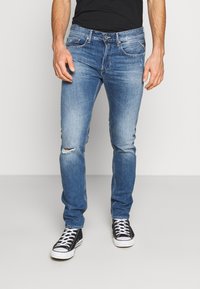 Hombre que lleva puestos unos vaqueros ajustados de color azul con un pequeño rasguño en la rodilla izquierda y unas zapatillas negras y blancas, de pie contra un fondo blanco.