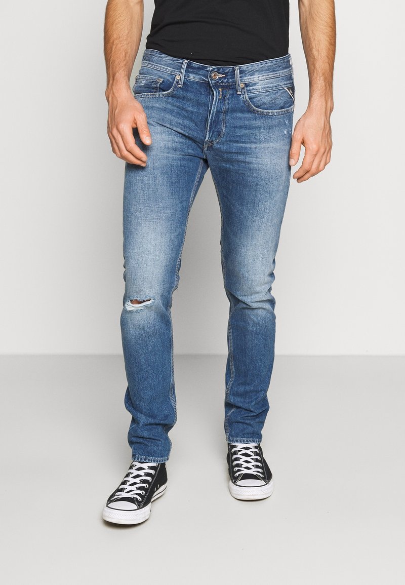 Hombre que lleva puestos unos vaqueros ajustados de color azul con un pequeño rasguño en la rodilla izquierda y unas zapatillas negras y blancas, de pie contra un fondo blanco.