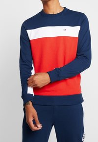 Marinblå sweatshirt med röda och vita horisontella ränder, rund halsringning och ribbad ärmslut. Har logotyp på bröstet. Tillverkad av bomullsblandat tyg.