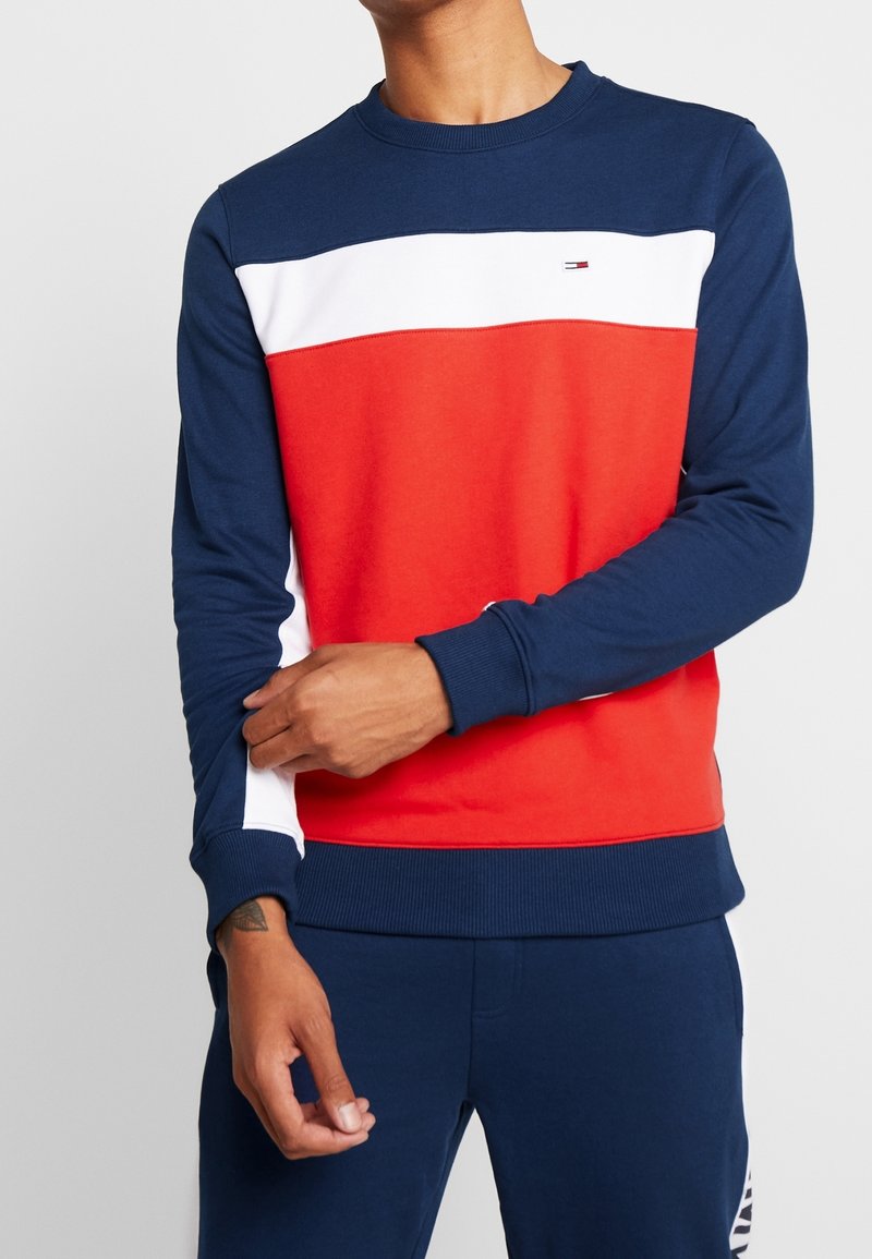 Marinblå sweatshirt med röda och vita horisontella ränder, rund halsringning och ribbad ärmslut. Har logotyp på bröstet. Tillverkad av bomullsblandat tyg.