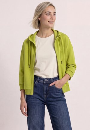 Młoda kobieta z krótkimi blond włosami w limonkowej bluzie na zamek, białej koszulce i niebieskich dżinsach, uśmiechająca się i patrząca na bok.