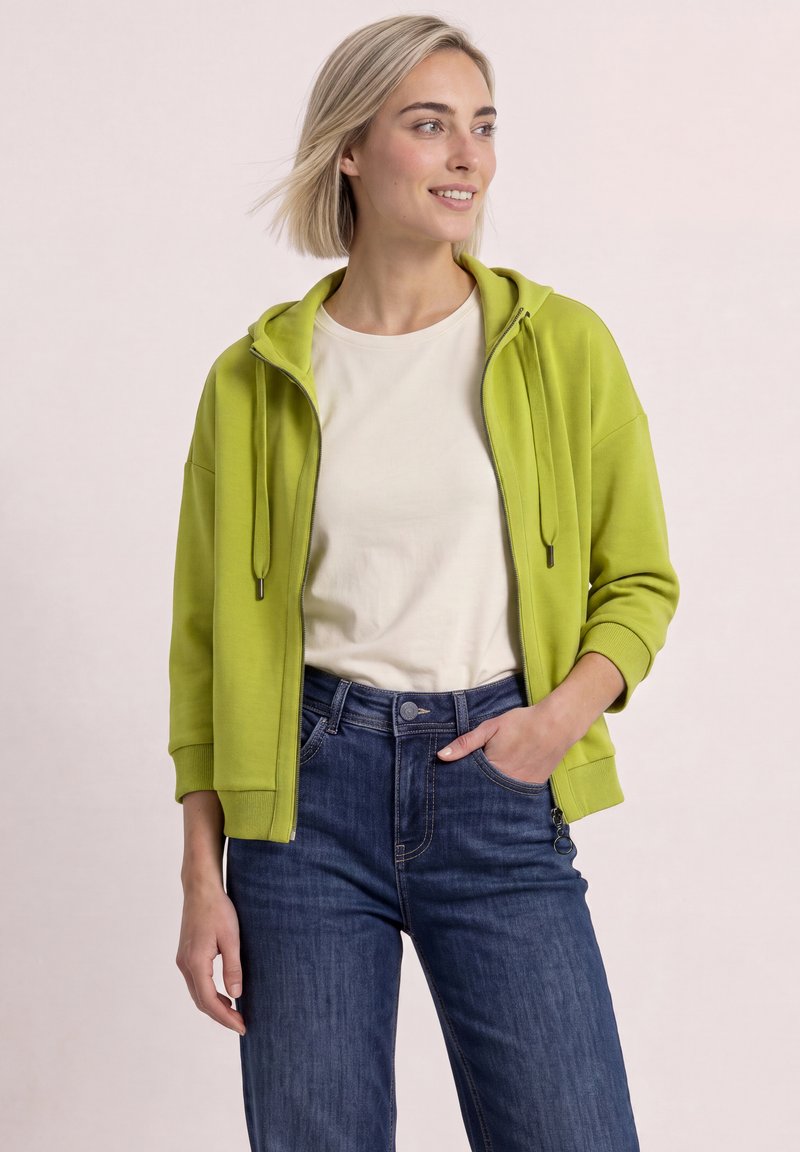 Jeune femme aux cheveux blonds courts portant un sweat à capuche zippé vert citron vert, un t-shirt blanc et un jean bleu, souriante et regardant sur le côté.