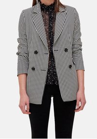 Svartvitt houndstoothblazer med dubbelknäppt design, spetsiga slag och svarta knappar, bärs över en genomskinlig svart blus med polkadots.