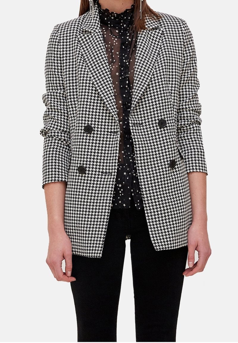 Svartvitt houndstoothblazer med dubbelknäppt design, spetsiga slag och svarta knappar, bärs över en genomskinlig svart blus med polkadots.