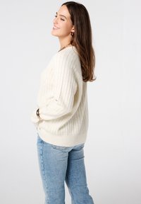 Romige geribbelde gebreide trui met een relaxte pasvorm, lange mouwen en een ronde halslijn, gecombineerd met lichtblauwe jeans.