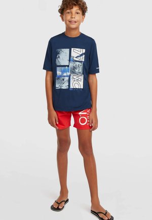 Dreng med krøllet hår iført en navyblå t-shirt med grafik, røde shorts med hvid tekst og sorte klipklappere, der står på en ensfarvet baggrund.