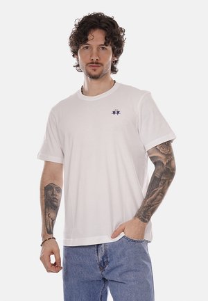 Uomo con capelli ricci, indossa una maglietta bianca e jeans blu, mostra tatuaggi dettagliati su entrambi gli avambracci, in piedi con una mano in tasca.