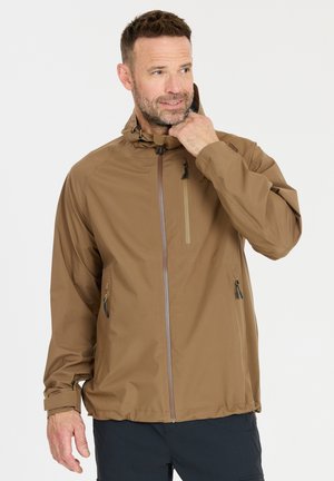 Mann, der die Kapuze einer braunen Jacke mit Reißverschluss und schwarzen Hosen gegen einen schlichten Hintergrund anpasst.