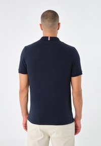 Námořnická modrá polo košile vyrobená z hladké látky, s klasickým límečkem, krátkými rukávy a pruhovaným akcentem na zadní straně límce.