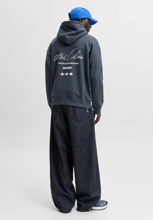 Persoon die een grijze hoodie draagt met witte tekst en sterren op de achterkant, brede donkere spijkerbroek, blauwe sneakers en een blauwe baseballpet, zijwaarts gekeerd.