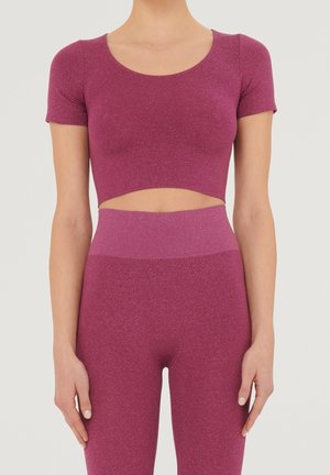 Femme portant un crop top magenta scintillant à manches courtes et des leggings taille haute assortis, debout devant un fond clair uni.